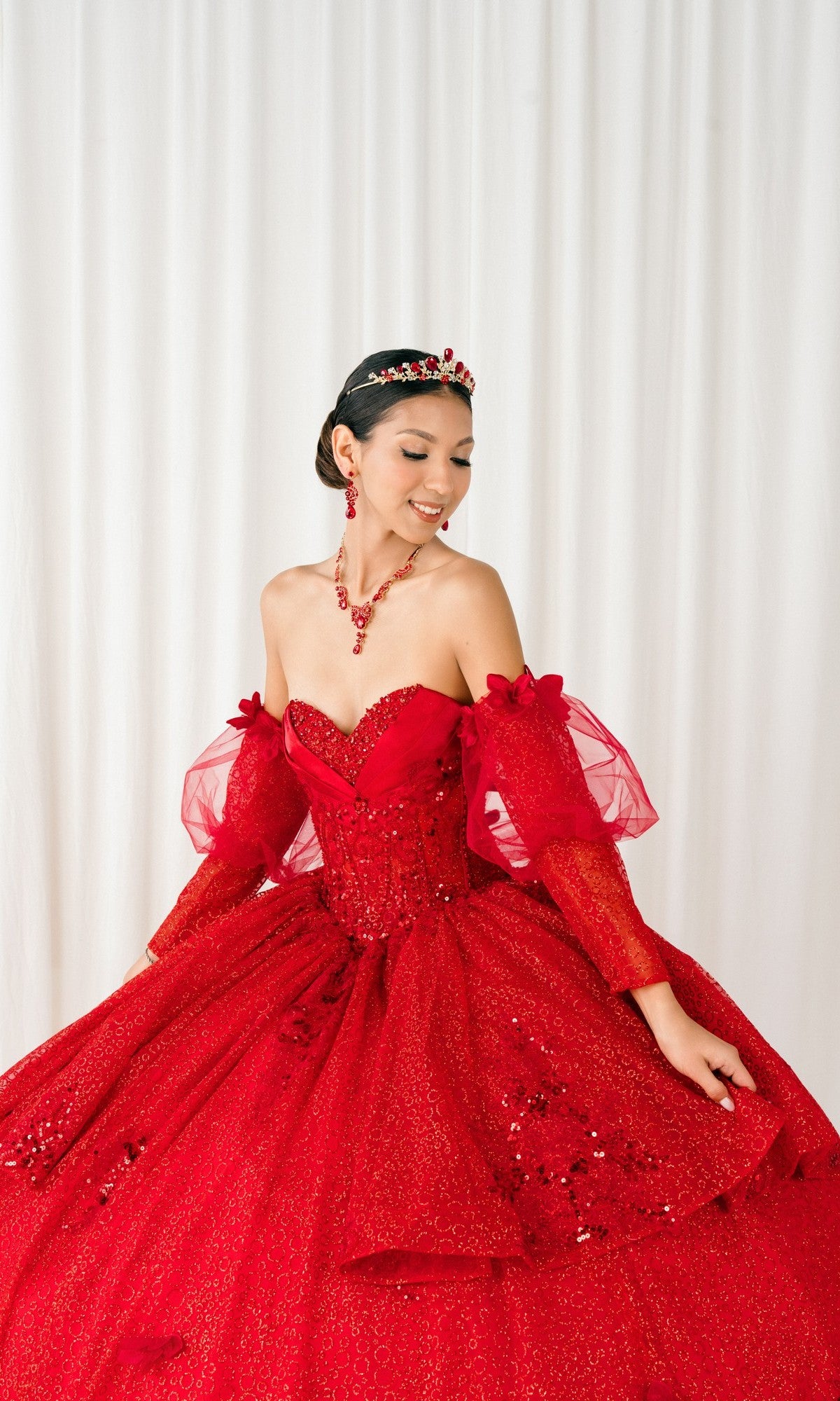 A woman wears the Quinceanera Dress 8385J By Cinderella Couture, a red off-the-shoulder gown with beaded details and puff sleeves, paired with matching jewelry and a tiara, standing before a white curtain backdrop.